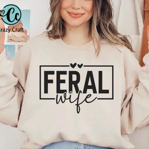 May include: A white sweatshirt with the text "FERAL wife" in black lettering inside a black rectangle. Two small hearts are above the rectangle.