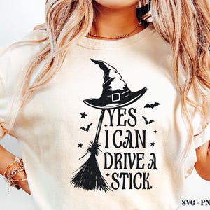 K&ouml;nnte beinhalten: Cremefarbenes T-Shirt mit schwarzem Hexenhut, Besen, Flederm&auml;usen und Sternen-Grafik. Der Text lautet "YES I CAN DRIVE A STICK." Das Crazy Craft-Logo befindet sich oben rechts.