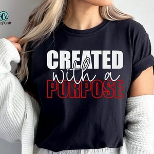 以下が含まれることがあります： 白と赤の文字で「CREATED with a PURPOSE」と書かれた黒いTシャツ。 シャツはクルーネックと半袖です。 Crazy Craftのロゴが左上にあります。