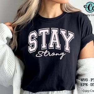 May include: Black t-shirt with the words "STAY Strong" in a pink, collegiate-style font. The shirt also has the text "Strong" in a cursive font. The shirt is a casual style.