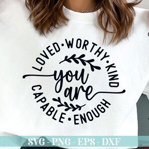 Pu&ograve; includere: Felpa bianca con un design circolare nero che dice "Loved-Worthy-Kind You Are Capable-Enough" con un piccolo disegno floreale su entrambi i lati del testo "You Are".