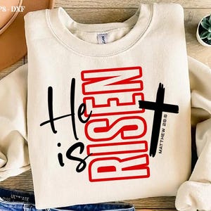 May include: Cream-colored sweatshirt with the text "He is Risen" in black and red, featuring a cross and the Bible verse Matthew 28:6. The design is centered on the front of the long-sleeved top.