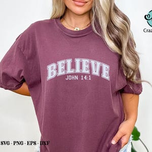 K&ouml;nnte beinhalten: Ein maroonfarbenes T-Shirt mit dem Wort "BELIEVE" in wei&szlig;en Buchstaben mit grauem Umriss. Der Text "JOHN 14:1" ist unter dem Wort "BELIEVE" gedruckt.