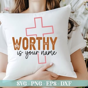 K&ouml;nnte beinhalten: Wei&szlig;es Kissen mit dem Text "WORTHY is your name" in Braun und Schwarz, mit einem rosa Kreuz. Das Kissen wird von einer Person gehalten, die ein wei&szlig;es Oberteil tr&auml;gt. Das Bild enth&auml;lt auch den Text "SVG - PNG - EPS - DXF".