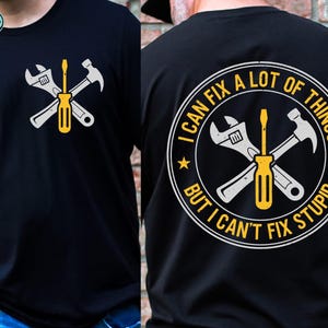 May include: Black t-shirt featuring a crossed wrench, screwdriver, and hammer graphic in white and yellow. The back of the shirt displays a circular design with the text "I CAN FIX A LOT OF THINGS BUT I CAN'T FIX STUPID."