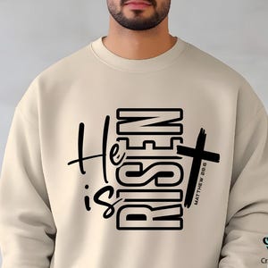 May include: Beige sweatshirt with the text "He is Risen" in black, featuring a cross graphic. The text "Matthew 28:6" is printed vertically on the cross. The sweatshirt has a crew neck and long sleeves.
