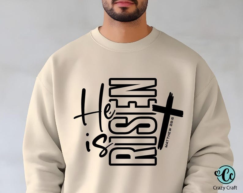 May include: Beige sweatshirt with the text "He is Risen" in black, featuring a cross graphic. The text "Matthew 28:6" is printed vertically on the cross. The sweatshirt has a crew neck and long sleeves.