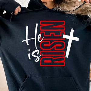May include: Black hooded sweatshirt with the text "He is Risen" in white script and red block letters. A white cross is to the right of the text. The sweatshirt has a front pocket and a drawstring hood.