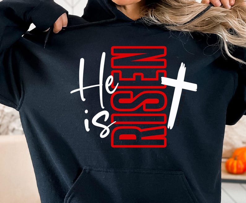 May include: Black hooded sweatshirt with the text "He is Risen" in white script and red block letters. A white cross is to the right of the text. The sweatshirt has a front pocket and a drawstring hood.