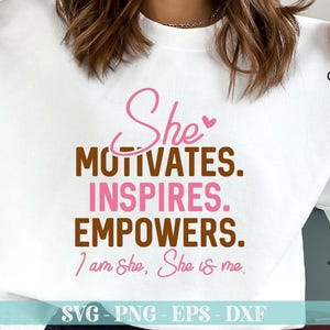 May include: A white t-shirt with a brown and pink graphic design that says "She motivates. Inspires. Empowers. I am She, She is me." The design is in a handwritten font. The t-shirt is on a white surface.