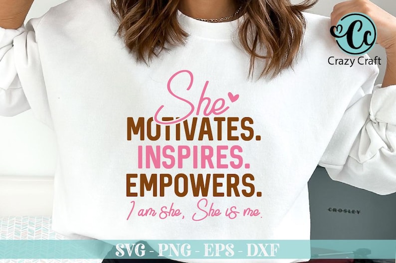 May include: A white t-shirt with a brown and pink graphic design that says "She motivates. Inspires. Empowers. I am She, She is me." The design is in a handwritten font. The t-shirt is on a white surface.