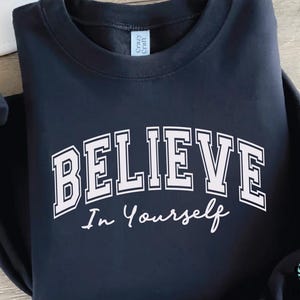 May include: A navy blue sweatshirt with the words "BELIEVE In Yourself" printed in white. The word "BELIEVE" is in a bold, collegiate-style font, and "In Yourself" is in a cursive font. The sweatshirt has a crew neck.