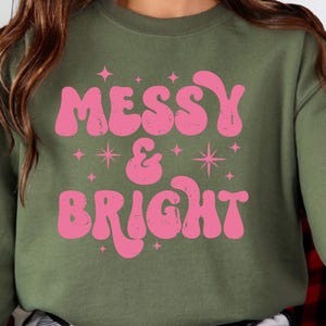 May include: Olive green sweatshirt with the words "MESSY & BRIGHT" in pink, retro-style lettering. The design includes star and sparkle graphics. The Crazy Craft logo is in the upper left corner.