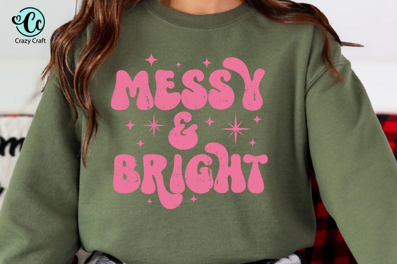 May include: Olive green sweatshirt with the words "MESSY & BRIGHT" in pink, retro-style lettering. The design includes star and sparkle graphics. The Crazy Craft logo is in the upper left corner.