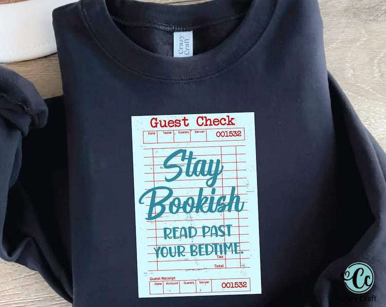 May include: Navy blue sweatshirt with a graphic of a guest check that reads "Stay Bookish, Read Past Your Bedtime." The design is in teal and red on a light blue background. The sweatshirt has a crew neck.