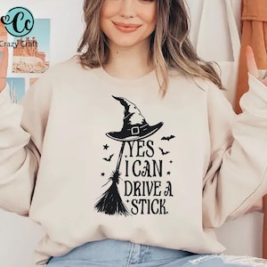 K&ouml;nnte beinhalten: Beiges Sweatshirt mit schwarzem Hexenhut, Besen und dem Text "YES I CAN DRIVE A STICK." Das Design beinhaltet Sterne und Flederm&auml;use, ideal f&uuml;r Halloween. Das Sweatshirt hat eine neutrale Farbe.