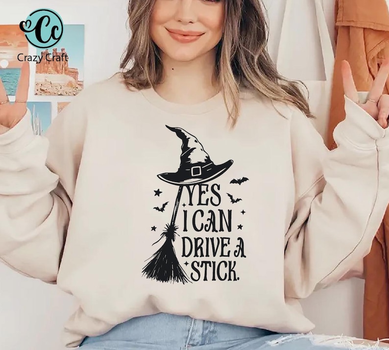 K&ouml;nnte beinhalten: Beiges Sweatshirt mit schwarzem Hexenhut, Besen und dem Text "YES I CAN DRIVE A STICK." Das Design beinhaltet Sterne und Flederm&auml;use, ideal f&uuml;r Halloween. Das Sweatshirt hat eine neutrale Farbe.