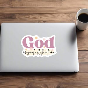 May include: A laptop with a sticker that reads "God is good all the time" in pink and black script. The laptop is on a wooden surface, next to a white coffee mug filled with coffee.