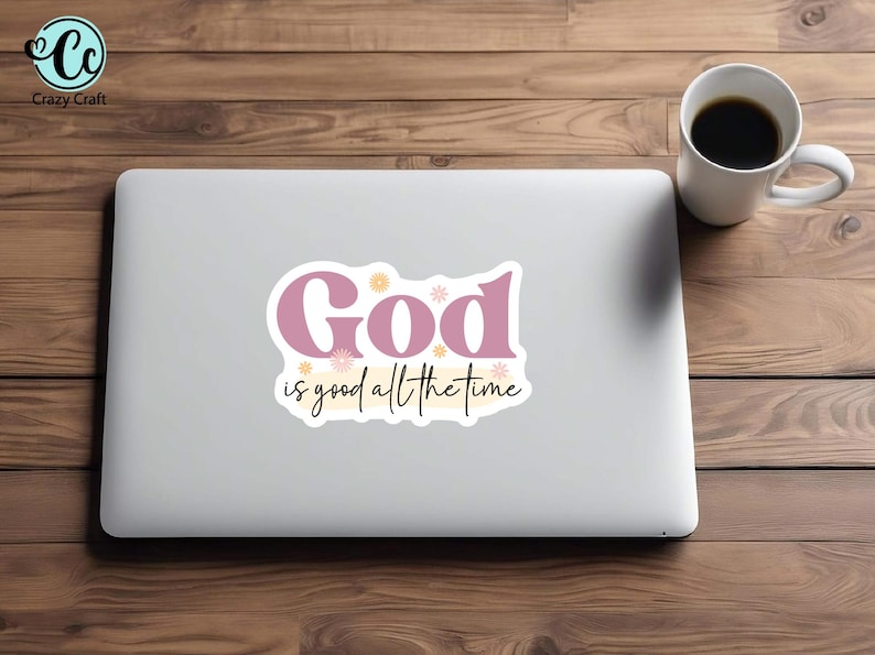 May include: A laptop with a sticker that reads "God is good all the time" in pink and black script. The laptop is on a wooden surface, next to a white coffee mug filled with coffee.