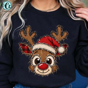 Puede incluir: Una sudadera negra de manga larga con un gráfico central de una cara de reno con grandes ojos marrones, una nariz roja y un gorro de Papá Noel rojo. El diseño tiene una apariencia texturizada de arpillera con bordes deshilachados.