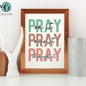 K&ouml;nnte beinhalten: Gerahmter Druck mit den Worten "PRAY on it, over it, through it" in T&uuml;rkis, Braun und Rosa. Eine wei&szlig;e dekorative Geweih und eine silberne Kerze befinden sich an den Seiten. Das Crazy Craft-Logo befindet sich oben links.