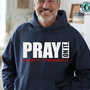 以下が含まれることがあります： 大きな白い文字で「PRAY ON IT」と書かれたネイビーブルーのフード付きスウェットシャツ。その下に、赤で「OVER IT. THROUGH IT.」と書かれています。