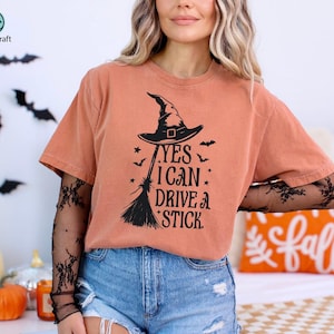 K&ouml;nnte beinhalten: Orangefarbenes T-Shirt mit schwarzem Hexenhut, Besen und dem Text "YES I CAN DRIVE A STICK". Das Shirt wird mit blauen Jeansshorts kombiniert. Halloween-Themenbekleidung.