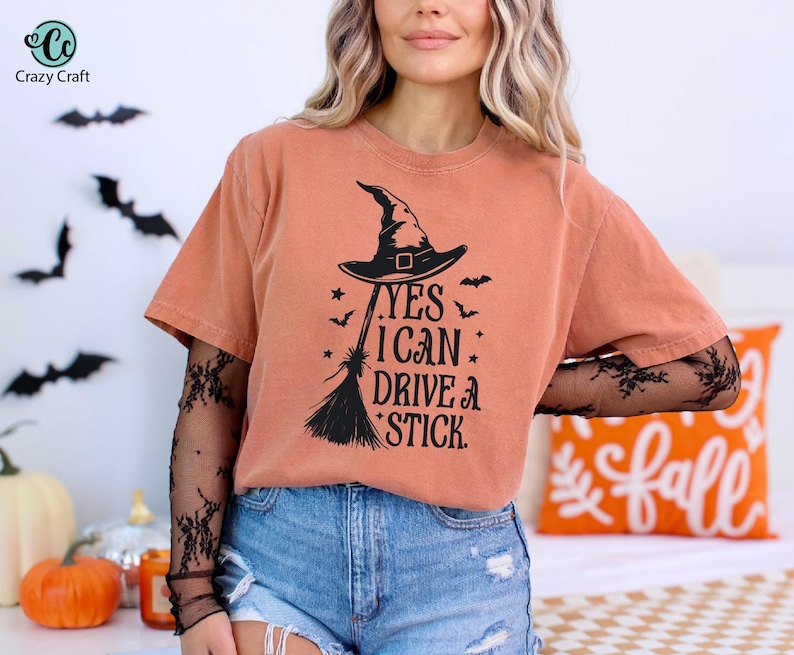 K&ouml;nnte beinhalten: Orangefarbenes T-Shirt mit schwarzem Hexenhut, Besen und dem Text "YES I CAN DRIVE A STICK". Das Shirt wird mit blauen Jeansshorts kombiniert. Halloween-Themenbekleidung.