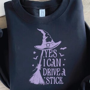 K&ouml;nnte beinhalten: Schwarzes Sweatshirt mit lila Hexenhut, Besen, Flederm&auml;usen und Sternen. Der Text lautet: "YES I CAN DRIVE A STICK."