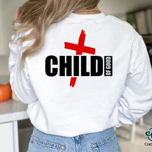 以下が含まれることがあります： 黒い文字で「CHILD OF GOOD」と赤い十字のデザインが施された白いスウェットシャツ。クルーネックと長袖です。Crazy Craftのロゴが右下に表示されています。
