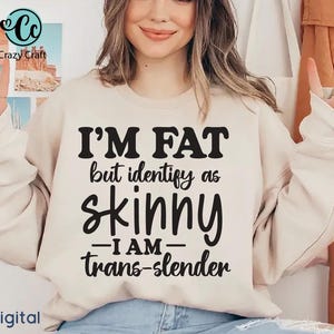 Pu&ograve; includere: Una felpa beige con la scritta "I'M FAT but identify as skinny - I AM - trans-slender" in nero. La felpa &egrave; indossata da una persona. La parola "Digital" si trova nell'angolo in basso a sinistra.