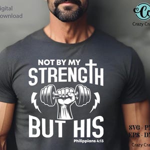 May include: Dark gray t-shirt with white text and graphics. The shirt features the words "NOT BY MY STRENGTH BUT HIS" with a dumbbell and cross design. The text "Philippians 4:13" is also included. The Crazy Craft logo is visible.