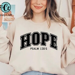K&ouml;nnte beinhalten: Ein cremefarbener Pullover mit dem Wort "HOPE" in schwarzen Blockbuchstaben mit schwarzem Umriss. Der Text "PSALM 130:5" ist in Schwarz unter dem Wort "HOPE" gedruckt.