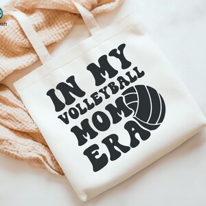 K&ouml;nnte beinhalten: Cremefarbene Tragetasche mit dem Text "IN MY VOLLEYBALL MOM ERA" in Schwarz, zusammen mit einer Volleyball-Grafik. Die Tasche hat zwei cremefarbene Tr&auml;ger und ist aus einem weichen Stoff gefertigt.
