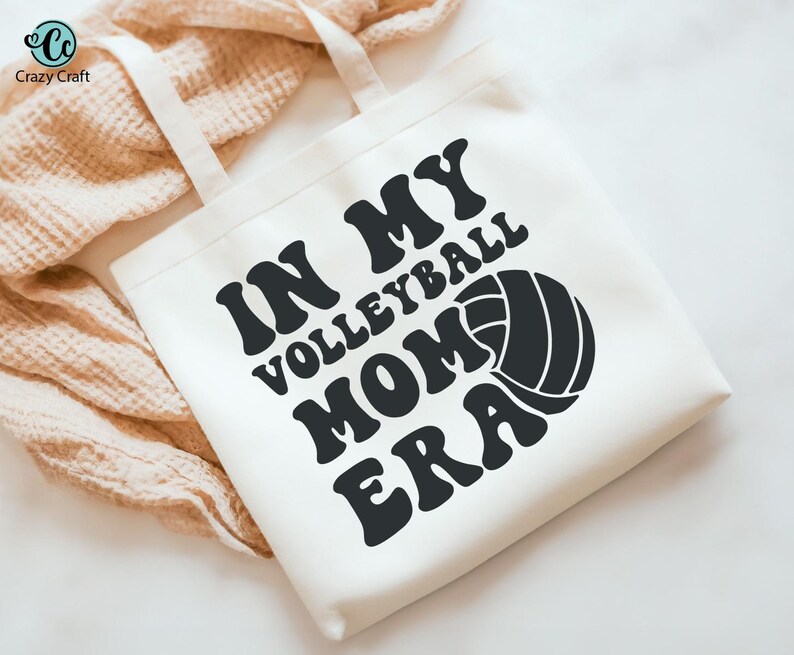 K&ouml;nnte beinhalten: Cremefarbene Tragetasche mit dem Text "IN MY VOLLEYBALL MOM ERA" in Schwarz, zusammen mit einer Volleyball-Grafik. Die Tasche hat zwei cremefarbene Tr&auml;ger und ist aus einem weichen Stoff gefertigt.