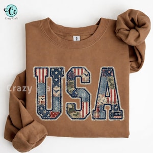 May include: A brown sweatshirt with the word "USA" in a patchwork design. The letters feature patterns of the American flag, including stars, stripes, and camouflage. The number "1776" is at the bottom right.