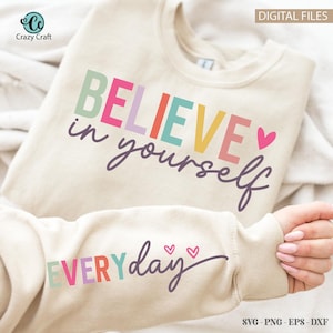 May include: A beige sweatshirt with the text "BELIEVE in yourself" in rainbow colours and a pink heart. The text "EVERYday" is also in rainbow colours on the sleeve.