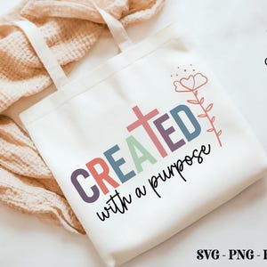 K&ouml;nnte beinhalten: Eine wei&szlig;e Tragetasche mit den Worten "CREATED with a purpose" in bunten Buchstaben und einer Kreuzgrafik. Ein Blumendesign befindet sich oben rechts. Die Tasche liegt auf einer hellen, strukturierten Oberfl&auml;che.