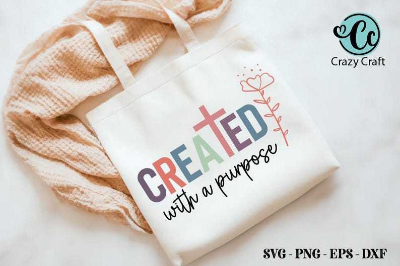 K&ouml;nnte beinhalten: Eine wei&szlig;e Tragetasche mit den Worten "CREATED with a purpose" in bunten Buchstaben und einer Kreuzgrafik. Ein Blumendesign befindet sich oben rechts. Die Tasche liegt auf einer hellen, strukturierten Oberfl&auml;che.