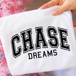 May include: A white t-shirt with the words "CHASE DREAMS" printed in bold, black, collegiate-style lettering. The shirt is held up by a hand, and the background includes pink and silver decorative elements.