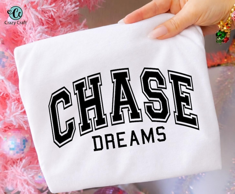 May include: A white t-shirt with the words "CHASE DREAMS" printed in bold, black, collegiate-style lettering. The shirt is held up by a hand, and the background includes pink and silver decorative elements.