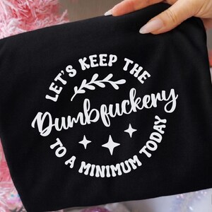 Pu&ograve; includere: T-shirt nero con la scritta bianca "LET'S KEEP THE Dumbfuckery TO A MINIMUM TODAY" in un design circolare. Il design include un rametto e quattro stelle. Il logo Crazy Craft si trova nell'angolo in alto a destra.