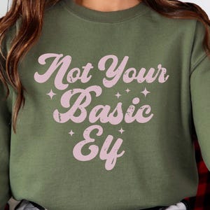 May include: An olive green sweatshirt with the text "Not Your Basic Elf" in a pink, retro font. The text is accented with small star shapes. The sweatshirt is a casual, comfortable style.
