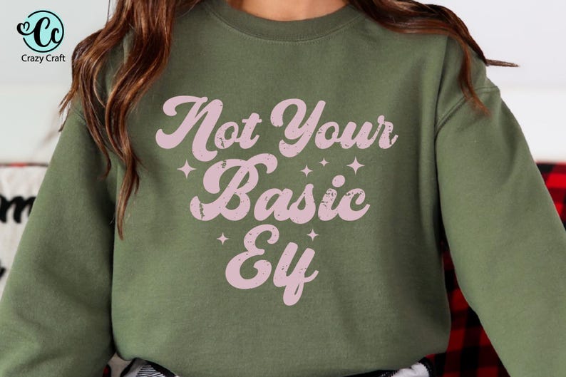 May include: An olive green sweatshirt with the text "Not Your Basic Elf" in a pink, retro font. The text is accented with small star shapes. The sweatshirt is a casual, comfortable style.