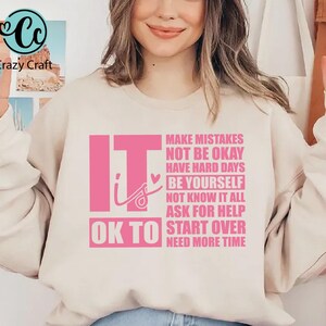 May include: A light beige sweatshirt with the text "It is OK to" in pink, followed by a list of phrases in pink, including "Make Mistakes", "Not Be Okay", "Have Hard Days", "Be Yourself", "Not Know It All", "Ask For Help", "Start Over", and "Need More Time".