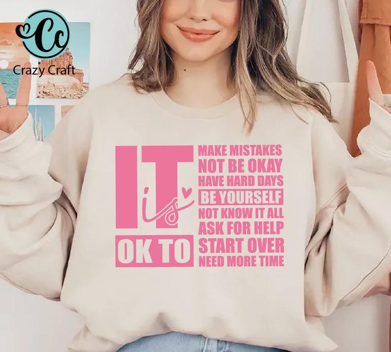 May include: A light beige sweatshirt with the text "It is OK to" in pink, followed by a list of phrases in pink, including "Make Mistakes", "Not Be Okay", "Have Hard Days", "Be Yourself", "Not Know It All", "Ask For Help", "Start Over", and "Need More Time".