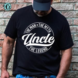 May include: Black t-shirt with the text "THE MAN THE MYTH Uncle THE LEGEND" in a white circular design. The shirt is short-sleeved and has a crew neck. The image is taken in front of a brick wall.