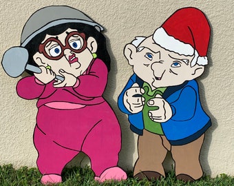 May include: A hand-painted wooden cutout of a couple dressed in winter clothing. The woman is wearing a pink outfit, a grey hat, and glasses. The man is wearing a blue jacket, brown pants, and a red Santa hat.