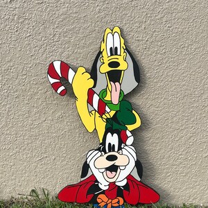 May include: A Christmas-themed wooden cutout of Goofy and Pluto. Goofy is wearing a red and white Santa hat and a red and white scarf. Pluto is wearing a green Santa hat and holding a red and white candy cane.