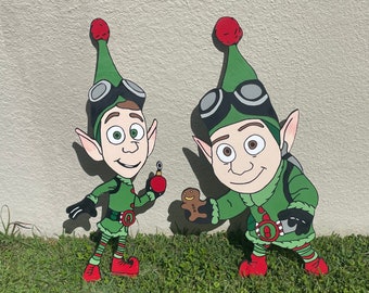 May include: Two green elf figures with red hats, goggles, and white faces. The elf on the left is holding a red ornament. The elf on the right is holding a gingerbread cookie.